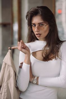 beautiful brunette girl in a sunglasses dressed in white blouse with a beige coat in hands. Stylish trendy fashion outlook.