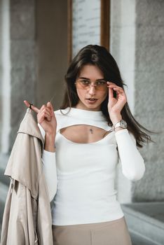 beautiful brunette girl in a sunglasses dressed in white blouse with a beige coat in hands. Stylish trendy fashion outlook.