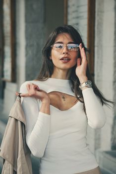 beautiful brunette girl in a sunglasses dressed in white blouse with a beige coat in hands. Stylish trendy fashion outlook.