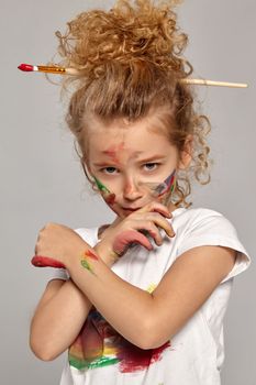 Beautiful little girl having a brush in her chic curly blond hair, wearing in a white t-shirt, has crossed her hands with a painted fingers, on a gray background.