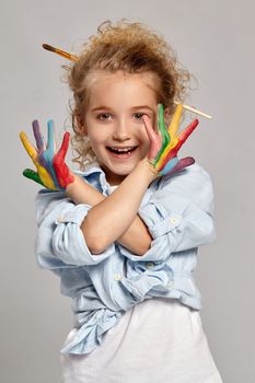 Pretty schoolgirl having a brush in her chic curly blond hair, wearing in a blue shirt and white t-shirt. She is showing her painted crossed hands and smiling at the camera, on a gray background.