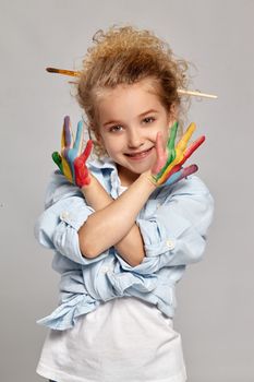 Cute schoolgirl having a brush in her chic curly blond hair, wearing in a blue shirt and white t-shirt. She crossed her painted arms, smiling and looking at the camera on a gray background.