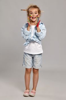 Cheerful child having a brush in her chic curly blond hair, wearing in a blue shirt and white t-shirt. She is posing with a painted hands and cheeks and looking wondered, on a gray background.