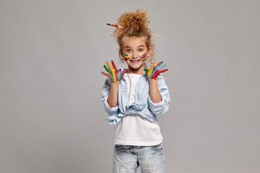 Lovely kid having a brush in her chic hairstyle, wearing in a blue shirt and white t-shirt. She raised her painted hands up, smiling and looking at the camera, on a gray background.
