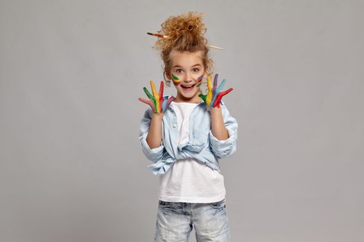 Lovely child having a brush in her chic hairstyle, wearing in a blue shirt and white t-shirt. She raised her painted hands up, looking at the camera and smiling widely, on a gray background.