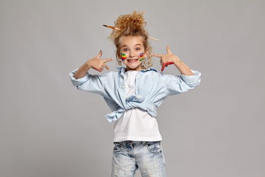 Lovely little lady having a brush in her chic curly blond hair, wearing in a blue shirt and white t-shirt. She has painted her cheeks and pointing on them, smiling at the camera on a gray background.