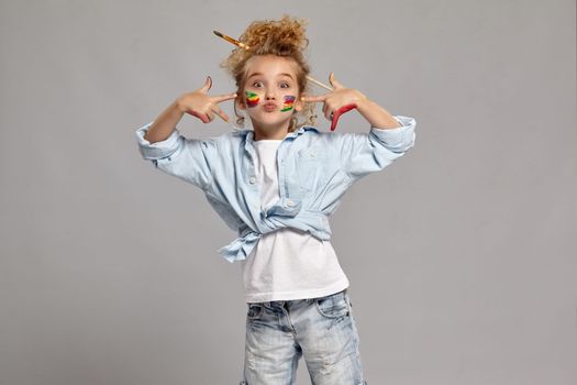 Pretty schoolgirl having a brush in her chic hairstyle, wearing in a blue shirt and white t-shirt. She has painted her cheeks and pointing on them, giving a kiss at the camera on a gray background.