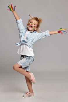 Beautiful schoolgirl a brush in her chic curly blond hair, wearing in a blue shirt and white t-shirt. She is posing with a painted hands and cheeks, smiling and looking very happy, on a gray background.