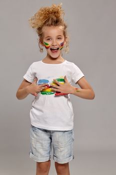 Wonderful kid having a brush in her lovely haircut, wearing in a white t-shirt. She is smearing her t-shirt, open her mouth and looking at the camera, on a gray background.