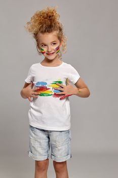 Wonderful child having a brush in her lovely haircut, wearing in a white t-shirt. She is smearing her t-shirt, smiling and looking away, on a gray background.