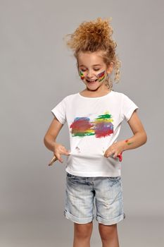 Beautiful little lady having a brush in her lovely haircut, wearing in a white smeared t-shirt. She is looking at it and smiling on a gray background.