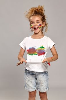 Beautiful kid having a brush in her lovely haircut, wearing in a white smeared t-shirt. She is holding it and looking satisfied, on a gray background.