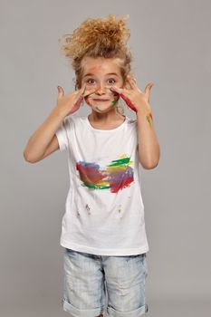Cute little girl having a brush in her lovely haircut, wearing in a white smeared t-shirt. She is smearing her face with paint and looking at the camera, on a gray background.