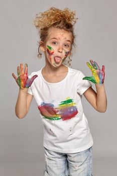 Cute child having a brush in her lovely haircut, wearing in a white smeared t-shirt. She is posing with a painted hands and cheeks and showing her tongue, on a gray background.