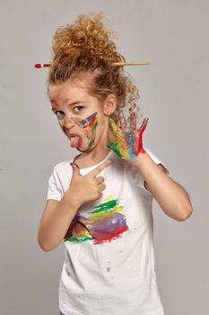 Beautiful little girl having a brush in her lovely haircut, wearing in a white smeared t-shirt. She is posing with a painted hands and cheeks, pointing on her hand and showing a tongue, on a gray background.