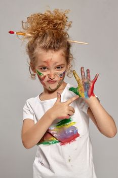 Beautiful child having a brush in her lovely haircut, wearing in a white smeared t-shirt. She is posing with a painted hands and cheeks, pointing on her hand and looking at the camera, on a gray background.