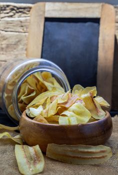 Homemade banana chips in wooden bowl and glass jar on sackcloth with vintage style. Banana dessert, Long banana chips, Empty place for text, Selective focus.