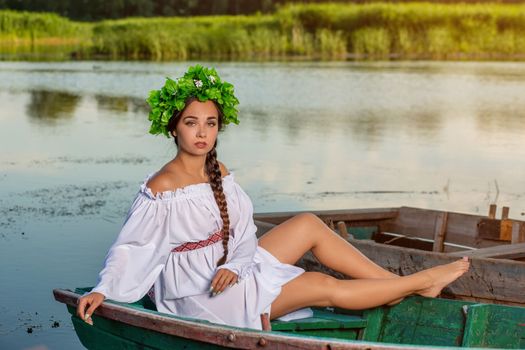 Young sexy woman on boat at sunset. The girl has a flower wreath on her head, relaxing and sailing on river. Beautiful body and face. Fantasy art photography. Concept of female beauty, rest in the village