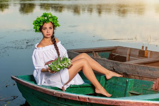 Young sexy woman on boat at sunset. The girl has a flower wreath on her head, relaxing and sailing on river. Beautiful body and face. Fantasy art photography. Concept of female beauty, rest in the village