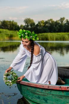 Young sexy woman on boat at sunset. The girl has a flower wreath on her head, relaxing and sailing on river. Beautiful body and face. Fantasy art photography. Concept of female beauty, rest in the village