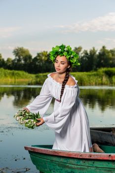 Young sexy woman on boat at sunset. The girl has a flower wreath on her head, relaxing and sailing on river. Beautiful body and face. Fantasy art photography. Concept of female beauty, rest in the village