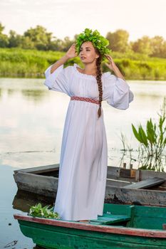 Young sexy woman on boat at sunset. The girl has a flower wreath on her head, relaxing and sailing on river. Beautiful body and face. Fantasy art photography. Concept of female beauty, rest in the village