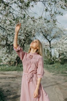 Blonde girl on a spring walk in the garden with cherry blossoms. Female portrait, close-up. A girl in a pink polka dot dress