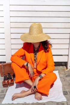 Stylish woman in an orange suit with a hat sits on a rug on a white striped background. On the hands are jewelry rings and bracelets, sandals and a bag stand side by side