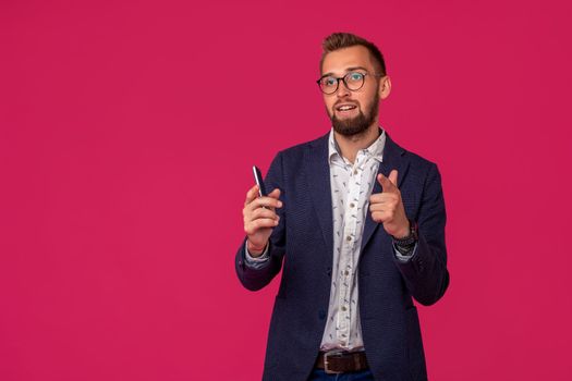 Studio shot of attractive brunette business man with glasses, in casual shirt, stylish black jacket holding a smartphone and smiling. Isolated pink background.