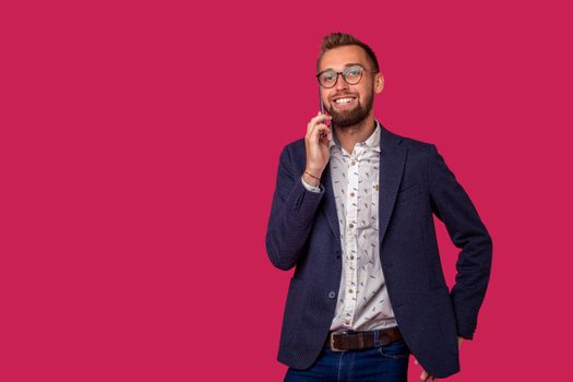 Studio shot of attractive brunette business man with glasses, in casual shirt, stylish black jacket talking on the cell phone and smiling. Isolated pink background.