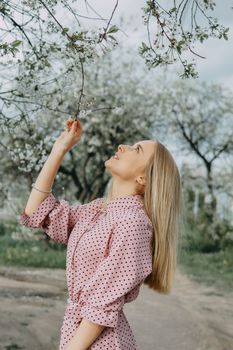 Blonde girl on a spring walk in the garden with cherry blossoms. Female portrait, close-up. A girl in a pink polka dot dress