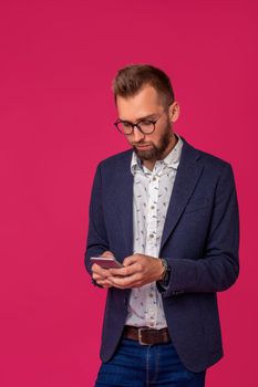 Studio shot of attractive brunette business man with glasses, in casual shirt, stylish black jacket holding a smartphone. Isolated pink background.