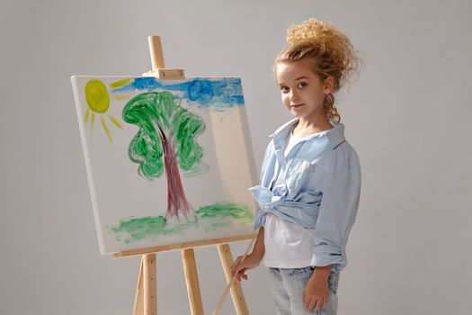 Charming school girl whith a curly blond hair, wearing in a blue shirt and white t-shirt is painting with a watercolor brush on an easel, looking at the camera and standing on a gray background.
