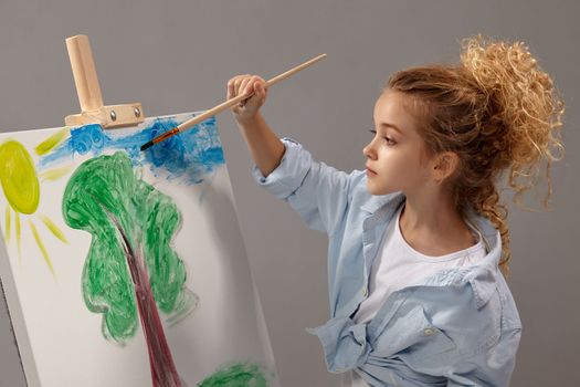 Amazing school girl whith a curly blond hair, wearing in a blue shirt and white t-shirt is painting with a watercolor brush on an easel, standing on a gray background.