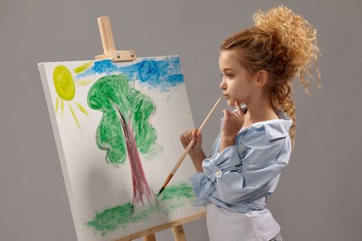 Thoughtful school girl whith a curly blond hair, wearing in a blue shirt and white t-shirt is painting with a watercolor brush on an easel, standing on a gray background.