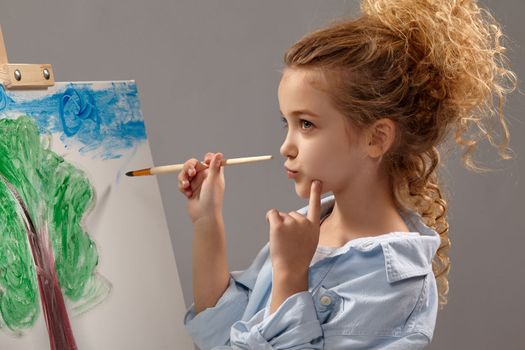 Thoughtful school girl whith a curly hair, wearing in a blue shirt and white t-shirt is painting with a watercolor brush on an easel, standing on a gray background.