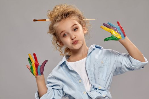 Beautiful little girl having a brush in her chic curly blond hair, wearing in a blue shirt and white t-shirt, is showing her painted fingers on a gray background.