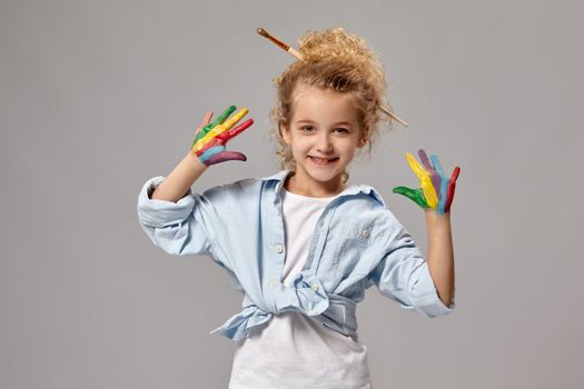 Beautiful little girl having a brush in her chic curly blond hair, wearing in a blue shirt and white t-shirt, with a painted fingers is posing on a gray background.