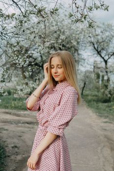 Blonde girl on a spring walk in the garden with cherry blossoms. Female portrait, close-up. A girl in a pink polka dot dress