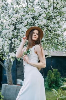 Beautiful young girl in white dress and hat in blooming Apple orchard. Blooming Apple trees with white flowers