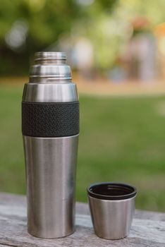 Stainless steel cup, mug with thermos stands on a wooden table in the forest on a green natural background. Thermos and mug with hot drink standing on wet wooden table after rain outdoors, close-up. Steam rises from thermo mug