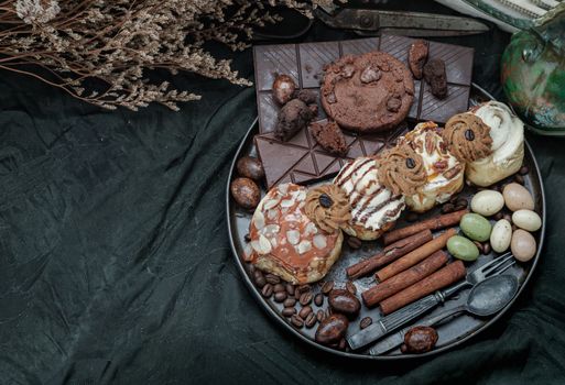 Assorted Cinnamon Rolls, Dark chocolate bars, cookies, pieces and balls served with black iron cutlery in black ceramic plate on dark background. Top view, Space for text, Selective