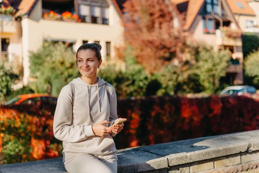 Young fashionable teenage girl with smartphone in park in autumn sitting at smiling. Trendy young woman in fall in park texting. Retouched, vibrant colors. Beautiful blonde teenage girl wearing casual modern autumn outfit sitting in park in autumn. Retouched, vibrant colors, brownish tones.