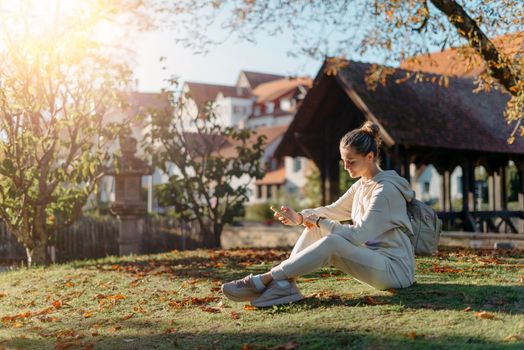 Young fashionable teenage girl with smartphone in park in autumn sitting at smiling. Trendy young woman in fall in park texting. Retouched, vibrant colors. Beautiful blonde teenage girl wearing casual modern autumn outfit sitting in park in autumn. Retouched, vibrant colors, brownish tones.