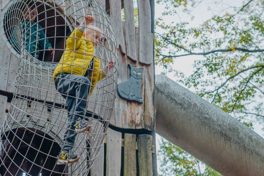 A child climbs up an alpine grid in a park on a playground on a hot summer day. children's playground in a public park, entertainment and recreation for children, mountaineering training. Child playing on outdoor playground. Kids play on school or kindergarten yard. Active kid on colorful slide and swing. Healthy summer activity for children. Little boy climbing outdoors.