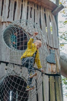 A child climbs up an alpine grid in a park on a playground on a hot summer day. children's playground in a public park, entertainment and recreation for children, mountaineering training. Child playing on outdoor playground. Kids play on school or kindergarten yard. Active kid on colorful slide and swing. Healthy summer activity for children. Little boy climbing outdoors.