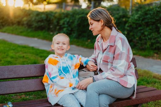 mother and son sit on a park bench in the rays of the setting sun. the concept of a family. Mother's Day. beautiful girl (mother) with a boy (son) in the park in the park are sitting on a bench at sunset.