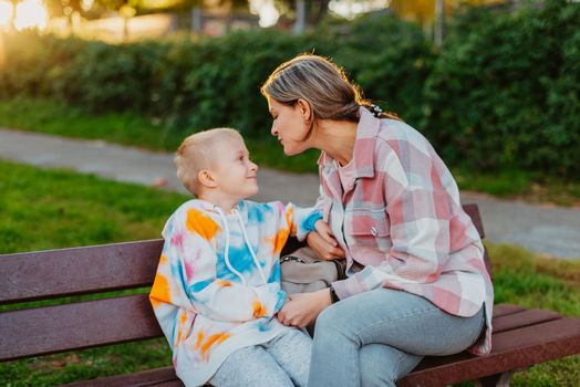 mother and son sit on a park bench in the rays of the setting sun. the concept of a family. Mother's Day. beautiful girl (mother) with a boy (son) in the park in the park are sitting on a bench at sunset.