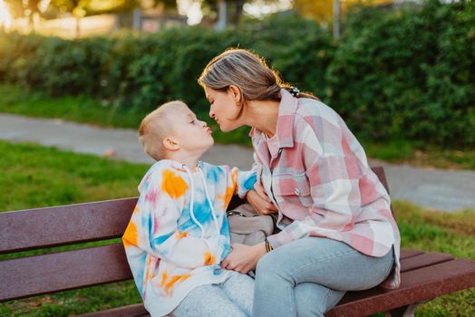 mother and son sit on a park bench in the rays of the setting sun. the concept of a family. Mother's Day. beautiful girl (mother) with a boy (son) in the park in the park are sitting on a bench at sunset.