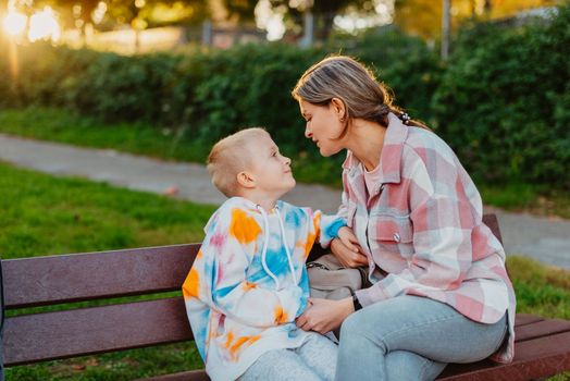 mother and son sit on a park bench in the rays of the setting sun. the concept of a family. Mother's Day. beautiful girl (mother) with a boy (son) in the park in the park are sitting on a bench at sunset.